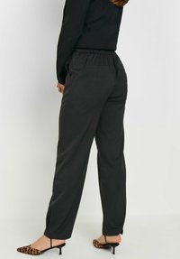Zwarte, elastische taillebroek met een relaxte pasvorm, voorzien van zijzakken en splitten aan de onderkant, gedragen met lederen schoenen met luipaardprint.