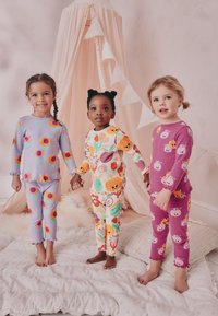 Drei Kinder tragen bunte Schlafanzüge: eines in Flieder mit Blumenmustern, eines in Creme mit Tiermotiven und eines in Pink mit Cartoonfiguren.