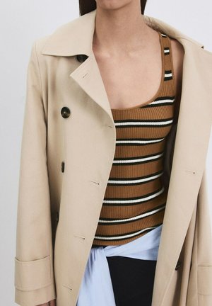 Persona che indossa un trench beige sopra una canotta a righe marrone, nero e bianco, con un capo azzurro legato in vita.