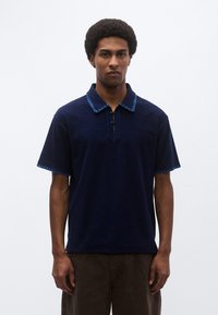 Μπλε σκούρο πόλο πουκάμισο με υφή σε ύφασμα, κοντά μανίκια και μπλε διακοσμητικό ράψιμο στο γιακά και το τελείωμα. Πέντε κουμπιά στη λωρίδα κουμπώματος.