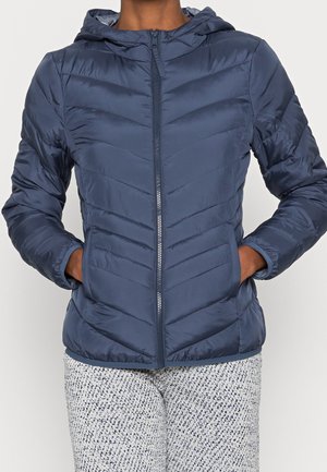 Personne portant une doudoune matelassée bleu marine avec capuche et fermeture éclair devant, mains dans les poches, et pantalon clair à motifs.