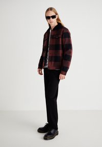 Red and black checkered jacket with black collar, worn over a gray shirt. Black pants and chunky black shoes complete the outfit.