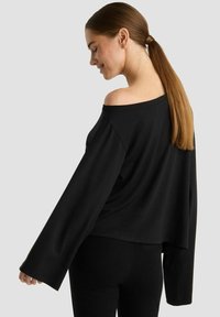 Schwarzes, kurzes Oberteil mit schulterfreiem Design, langen Ärmeln und glatter Textur. Von der Seite an einer Person präsentiert.