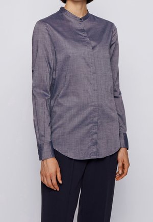 Žena v šedé košili s dlouhými rukávy a manžetovými knoflíky s mandlovým límečkem, v tmavých kalhotách, stojící na jednobarevném pozadí.