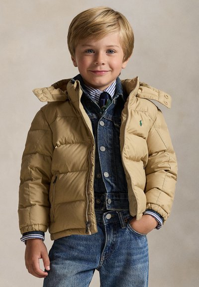 Blonder Junge trägt eine beige Steppjacke über einer Jeansjacke, gestreiftes Hemd, Krawatte und blaue Jeans, lächelt mit einer Hand in der Tasche.