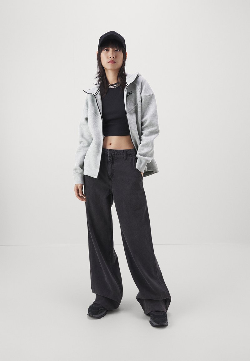 Grauer zweifarbiger Sweatshirt, schwarzes Crop-Top, weit geschnittene schwarze Hose und schwarze Turnschuhe. Schwarze Kappe mit Logo und Kettenhalskette.