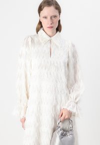 Robe blanche avec des franges texturées en zigzag, dotée d'un col, d'une fermeture à boutons et de manches longues. Sac pochette gris.