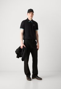 Czarny polo z akcentami w kolorze turkusowym przy kołnierzy, czarne spodnie i czarne buty. Model trzyma czarną kurtkę. Prosty design i dopasowany krój.