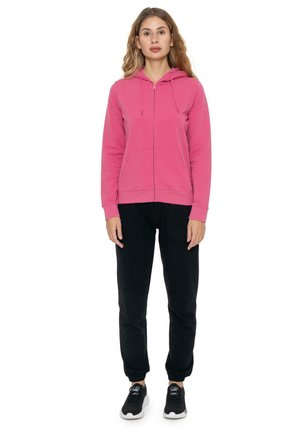 Une femme se tient devant un fond blanc, vêtue d’un sweat à capuche rose zippé, d’un pantalon de jogging noir et de baskets noires.