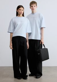 Φαρδιά, ανοιχτό μπλε μπλουζάκια με διακριτικό λογότυπο, σε συνδυασμό με μαύρο παντελόνι φαρδιάς γραμμής. Ένα άτομο κρατάει μια μαύρη, τετράγωνη τσάντα.