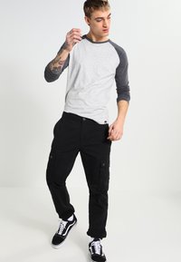 Maglietta a maniche lunghe raglan grigia e nera abbinata a pantaloni cargo neri e sneakers nere. Presenta tatuaggi sul braccio.