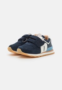 Baskets bleu marine avec des matériaux en daim et en maille, dotées de bandes Velcro, d'un accent blanc, et d'une semelle extérieure beige et bleue.