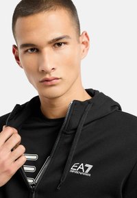 Sudadera con cremallera negra de tejido texturizado. Presenta una capucha con cordón y un logo blanco en relieve en el pecho. Llevada sobre una camiseta negra.