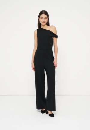 Nő fekete egyrészes, egyvállas overallt és fekete magas sarkú cipőt viselve, egyszínű fehér háttér előtt áll.