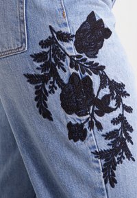 Svetle modré denimové džíny s jemným černým květinovým vyšíváním na stehně, s podrobnými listy a texturovaným prošíváním.
