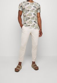 Kortärmad, rundhalsad skjorta med ett tropiskt bladmönster i grönt, rosa och blått. Matchad med krämfärgade avsmalnande byxor och bruna sandaler.