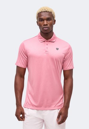 Jeune homme aux cheveux blonds bouclés, portant un polo rose clair à manches courtes et un pantalon blanc, debout devant un fond uni.