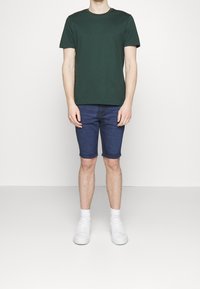 Osoba stojí na jednobarevném bílém pozadí, má na sobě zelené tričko, džínové šortky a bílé tenisky s kotníkovými ponožkami.