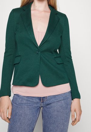 Kvinde iført en tætsiddende mørkegrøn blazer over en lys rosa bluse og blå jeans, stående mod en ensfarvet lys baggrund.