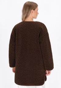 Giacca in pile marrone e morbido, con una vestibilità comoda, spalle scese e orlo arrotondato. La texture soffice e le maniche lunghe aumentano il calore.