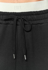 Zwarte trainingsbroek met een tulband taille, voorzien van zilveren aglets. De tailleband is een lichte kleur met subtiele branding. Zachte stoftextuur.