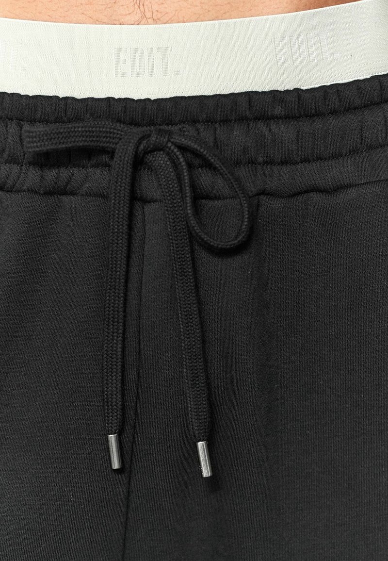 Zwarte trainingsbroek met een tulband taille, voorzien van zilveren aglets. De tailleband is een lichte kleur met subtiele branding. Zachte stoftextuur.