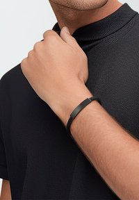 Black braided bracelet with a sleek, matte rectangular accent. Worn on a wrist, close-up against a textured black shirt.