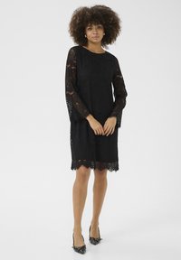 Abito in pizzo nero con maniche lunghe, fantasie floreali, orlo scallopato e una vestibilità rilassata, abbinate a scarpe nere con punta a stiletto.