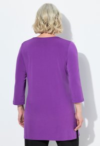 Fialová tunika s třičtvrtečními rukávy, hladkou texturou, kulatým výstřihem a délkou k bokům, v kombinaci s černými kalhotami.