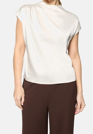 Mujer con blusa sin mangas de satén marfil y pantalones marrón oscuro, de pie frente a un fondo claro.
