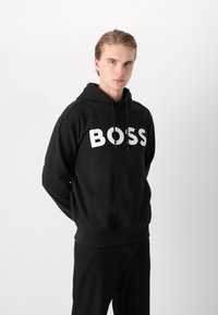 Juoda gobtuvė su dideliu baltu "BOSS" logotipu priekyje, turinti kengūrinę kišenę ir raištį ant gobtuvo. Lygi medžiagos tekstūra.