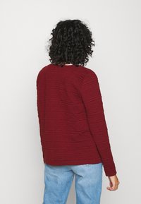 Suéter burdeos texturizado de corte relajado, con sutiles líneas horizontales y mangas largas, combinado con jeans de color azul claro.