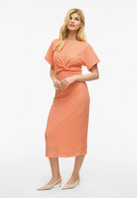 Robe jusqu'aux genoux en tissu texturé corail, avec un détail de taille torsadé et des manches courtes. Assortie à des talons beiges à bout pointu.