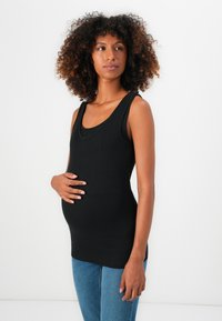 Top de tirantes anchos de canalé negro, diseño ajustado y escote redondeado, combinado con jeans de mezclilla azules. El sujeto está de pie con las manos en la barriga.