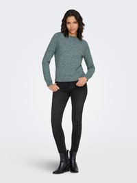 Roheline heegeldatud kampsun ümmarguse kaeluse ja pikkade varrukatega, kombineeritud mustade kitsaste teksade ja pahkluu saapadega. Sile tekstuur, keha järgnev lõige.