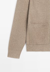 Manica e parte anteriore di maglione beige lavorato a maglia con tasca a toppa quadrata, polsini e orlo a coste.