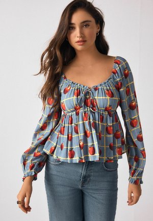 Floral blouse with puff sleeves, featuring a blue base and red apple pattern, and a cinched waist with a tie detail. Paired with denim jeans.