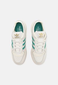 Vita Adidas-sneakers med teal-ränder, perforerat tåområde, vita snörningar och guld Adidas-logotyp på tungan, visade från ovan.