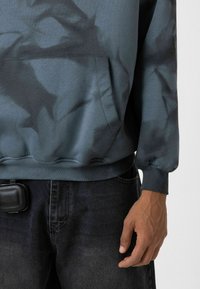 Een hoodie in gedempt teal met een donker, abstract patroon; voorzien van een voorzak, ribgebreide manchetten en een losse pasvorm. Draagt met donkere spijkerbroeken.