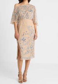 Beżowa sukienka na ramiączkach z haftem w srebrne kwiaty, przezroczysta nakładka, do kolan, z bocznym rozcięciem, w zestawieniu ze złotymi sandałami na paskach.