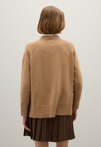 Beige Strickpullover mit hochgeschlossenem Kragen, geripptem Saum und überschnittenen Schultern, getragen über einem karierten Rock in verschiedenen Grüntönen und Braun.