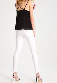 Camicetta nera con sottili spalline incrociate, abbinata a jeans bianchi aderenti e sandali con tacco argentati, che mostrano un aspetto elegante e contemporaneo.