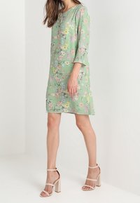 Grön blommig klänning med trekvartslånga ärmar, prydd med ett lätt mönster av rosa och gula blommor, i kombination med beige klacksandaler med blockklack.