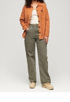 Frau trägt ein orangefarbenes Hemd über einem weißen Tanktop, olivfarbene Cargohose und beige Stiefel, steht mit einer Hand in der Tasche.