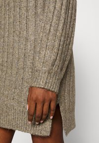 Gros plan sur la manche d'une robe pull en maille côtelée marron avec une fente latérale, une main à la peau foncée reposant contre le tissu.