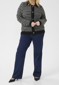 Schwarz-weißer, gemusterter Cardigan mit kragenförmigem Ausschnitt, Vorderknöpfen und gerippten Bündchen, kombiniert mit dunkelblauen Jeans im geraden Schnitt.