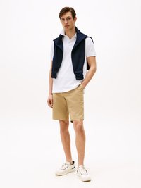 Witte polo, marineblauwe mouwloze sweater over de schouders, beige shorts en witte sneakers met zwarte accenten op een effen achtergrond.
