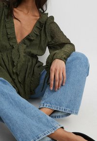 Blouse texturée vert olive avec un col et des manches à volants, associée à un jean flared bleu clair avec un ourlet brut.