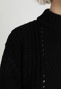 Pull noir en tricot avec un col montant, présentant un tissu texturé et un design de points partiellement ouverts sur le côté.