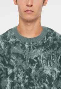 Adīta džemperis mīkstā, pūkainā audumā ar marmora zaļu rakstu. Tam ir apaļa kakla izgriezuma forma un teksturēta virsma.